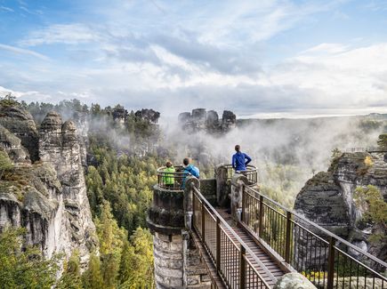 Eine Familie steht auf einer vorgelagerten Aussichtsplattform der Basteibrücke und schaut sich die Sandsteinformationen an. 