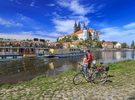 A couple standing with their bike on the Elbe Cycle Route in Meisen and looking out over the Elbe, where a steamship is moored, to Meissen's Albrechtsburg Castle. 