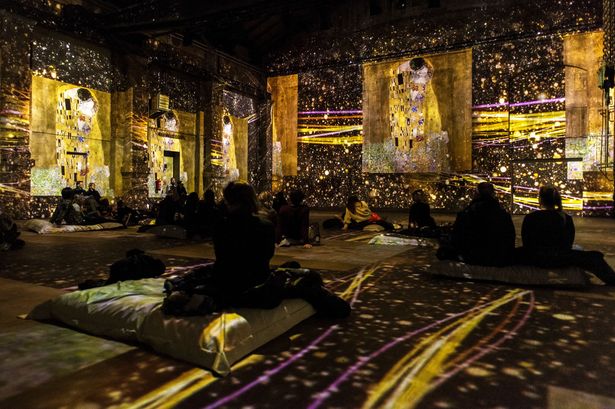 Besucher schauen sich auf dem Boden sitzend ein goldenes Kunstwerk von Gustav Klimt an, dass in einer multisensorischen Ausstellung den ganzen Raum ausfüllt