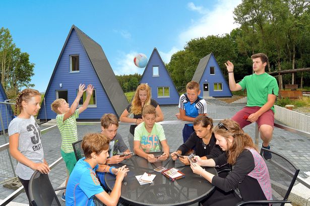 Kinder sitzen an einem Tisch und spielen Karten. Andere Kinder spielen Ball. Im Hintergrund sind die blauen Finnhütten des Schullandheims Limbach.